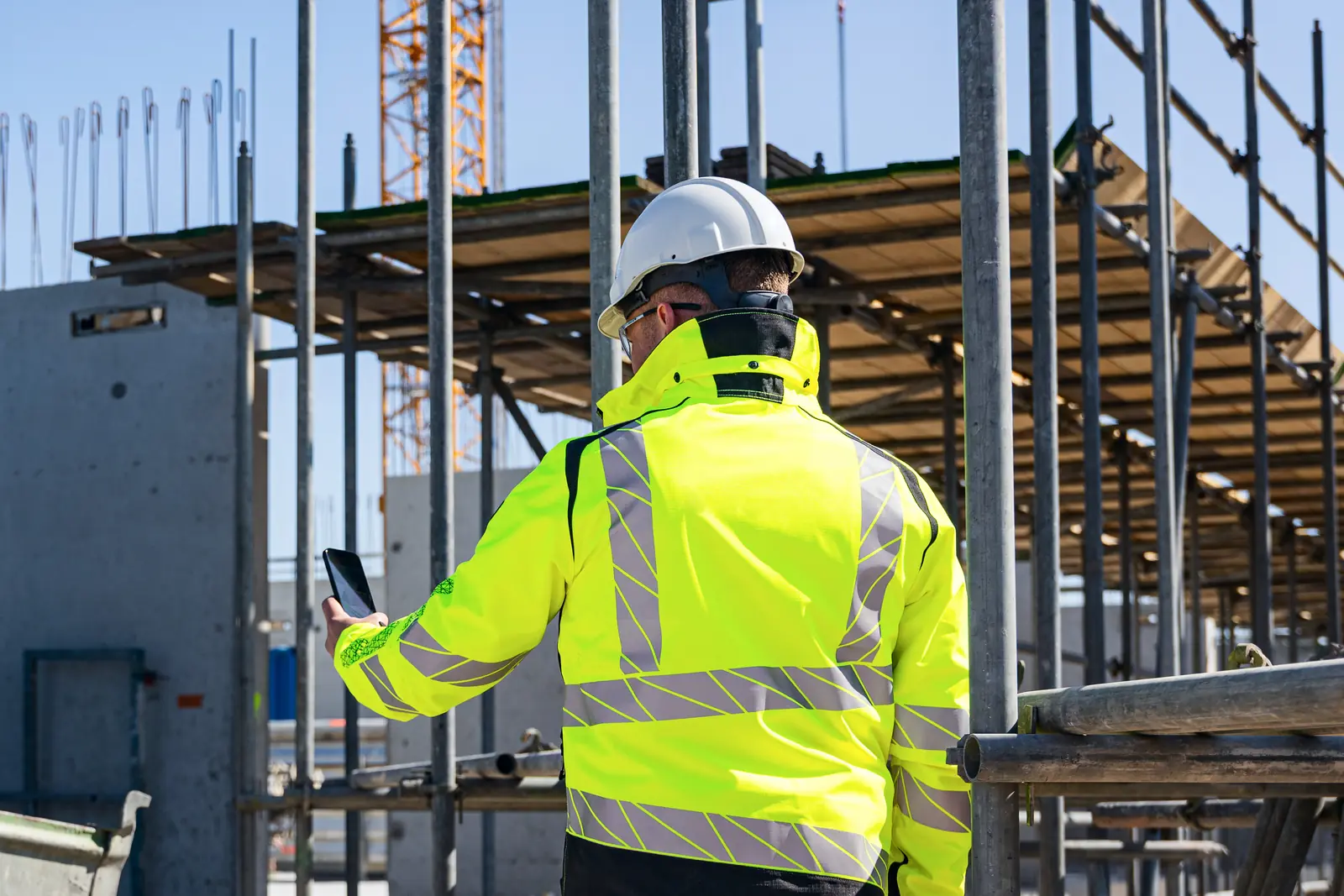 Een bouwplaats met steigers, betonwanden en een torenkraan. Een medewerker met een witte helm en een fluogele werkjas staat met zijn rug naar ons toe en heeft zijn telefoon in zijn hand.